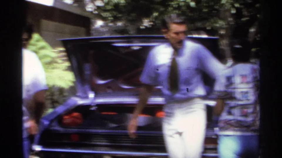 A man wearing a shirt and tie moves past the open trunk while another person stands at the edge of the frame during the family packing scene.