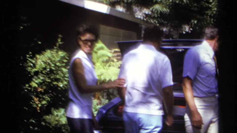 A smiling woman in sunglasses stands near two men at the rear of the car as the open trunk remains visible in the 1974 La Paz family driveway scene.