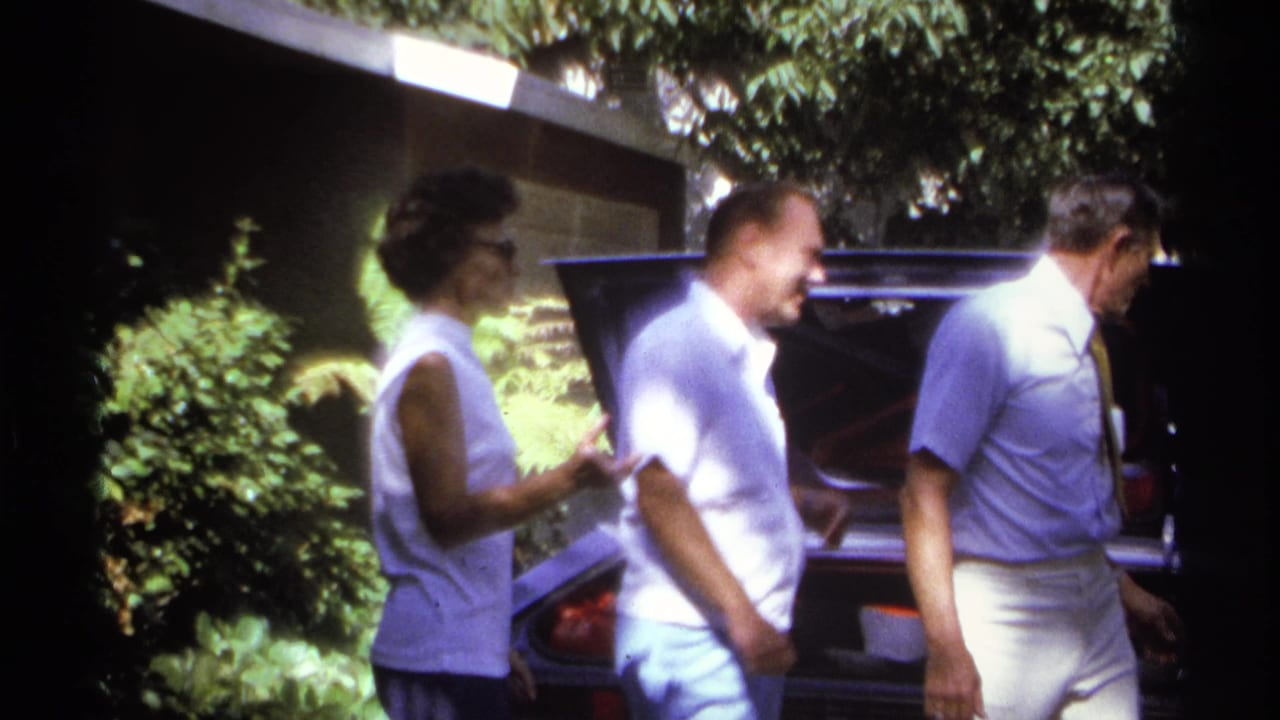 1974 La Paz home-movie frame showing adults beside an open car trunk under bright garden light.