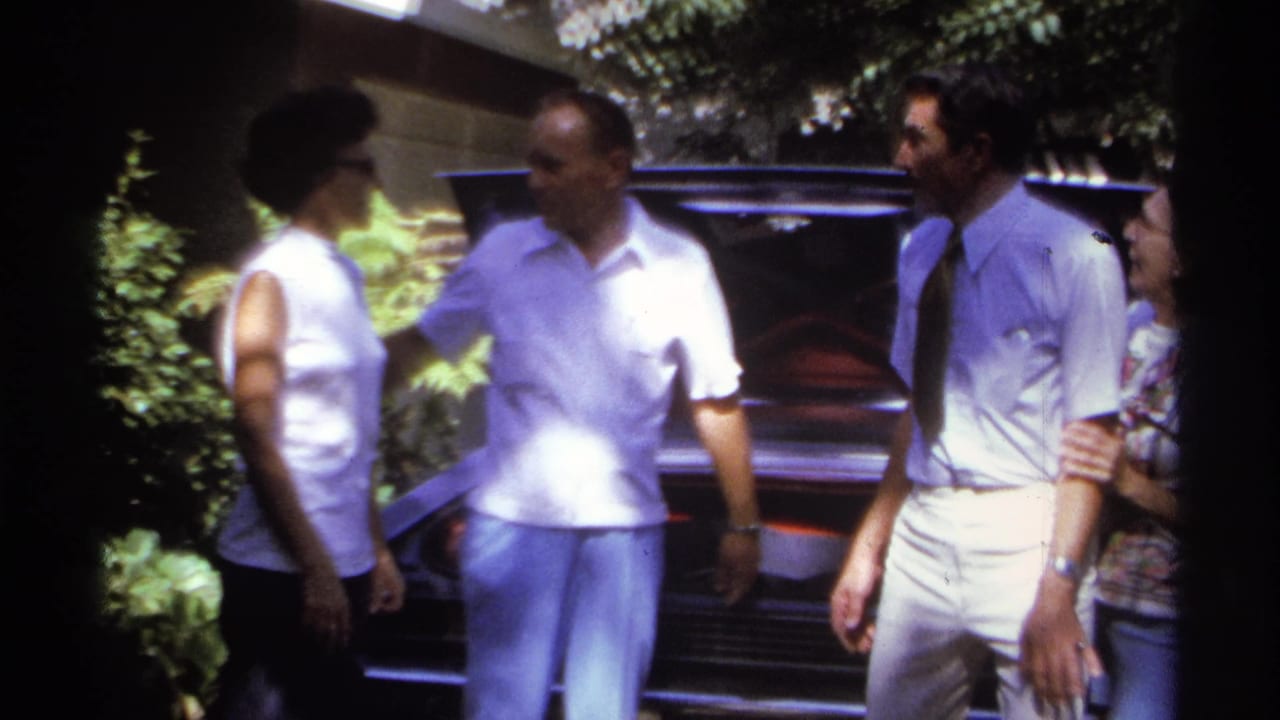 Adults converse beside an open car trunk, with trees and shrubs behind them.