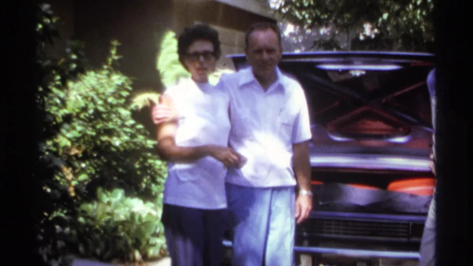 1974 La Paz home-movie still of a woman and man standing close together in front of an open car trunk.