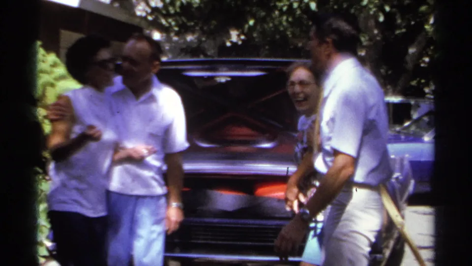 1974 La Paz home-movie frame showing adults laughing as one person gives a cheek kiss beside the open trunk.