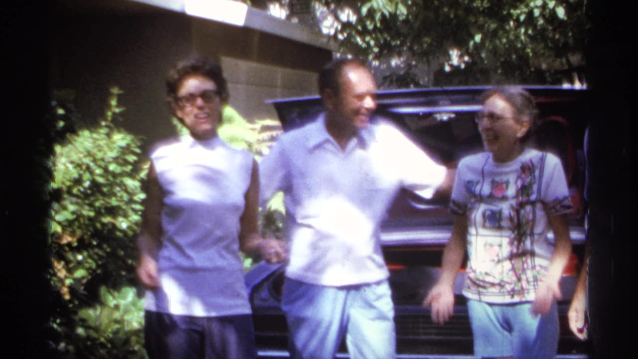Three adults smile and walk or pose in front of a car.