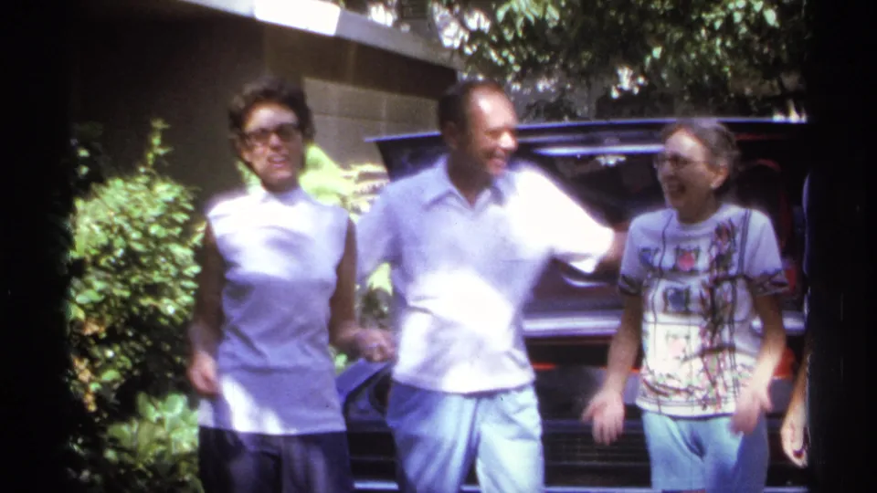 Three adults smile and move together near a car during a 1974 La Paz home-movie moment, based on metadata.