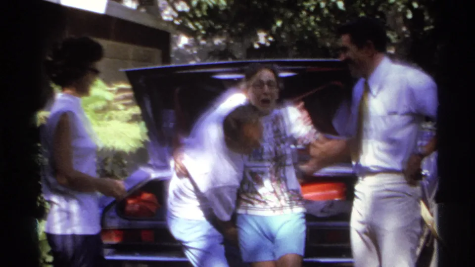 Adults laugh and lean toward one another beside an open car rear in a sunlit 1974 La Paz home-movie scene.
