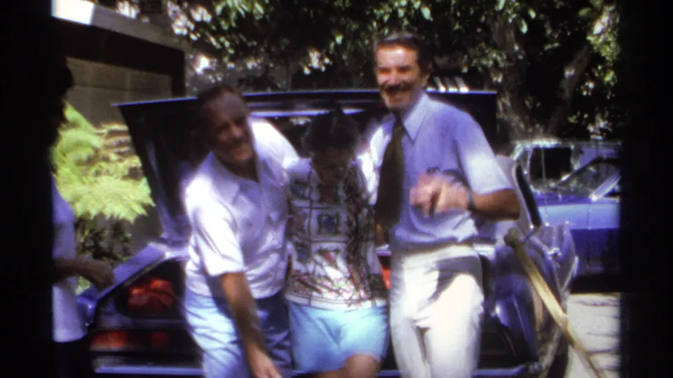 Three adults laugh, lean together, and gesture toward the camera beside a car in metadata-supported 1974 La Paz footage.
