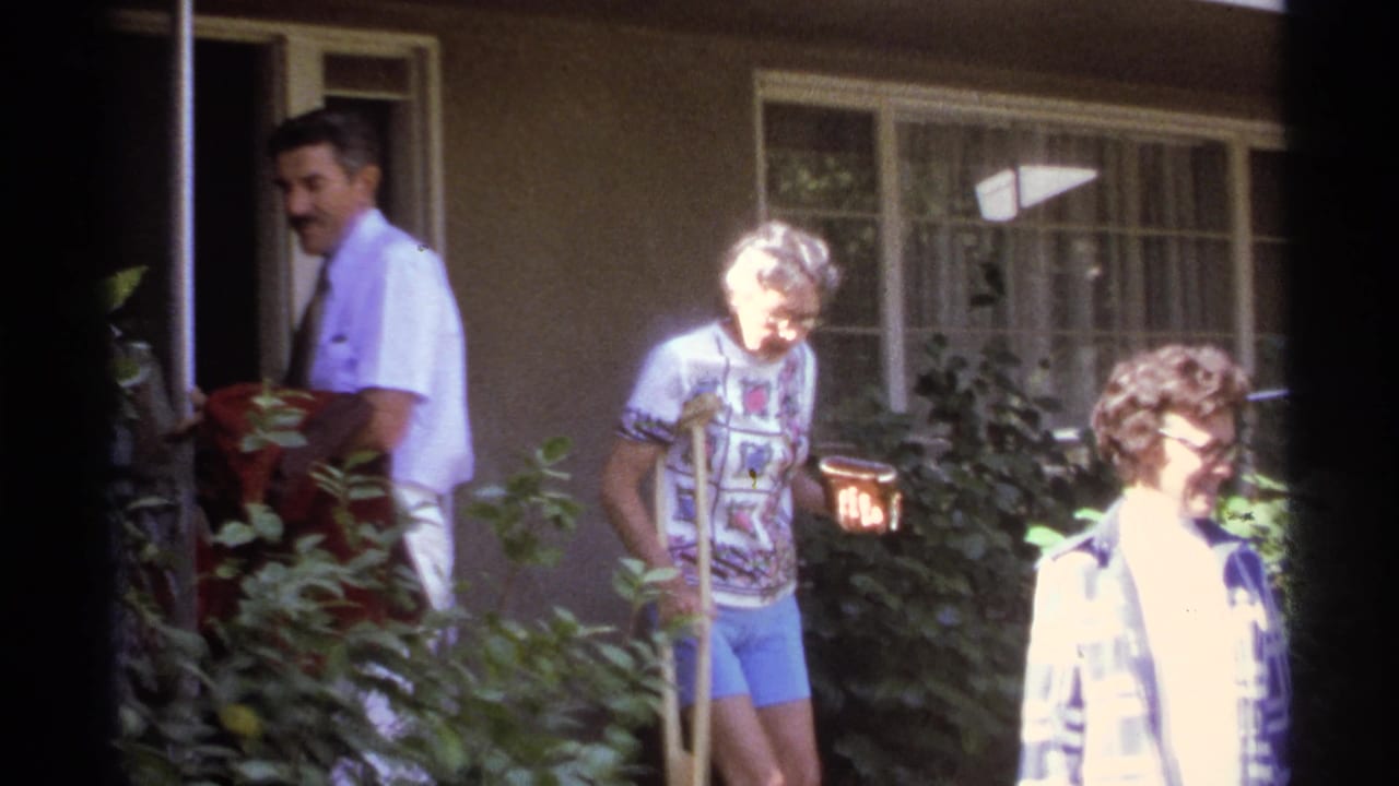 Older adults outside a house, with one woman using crutches near garden shrubs.