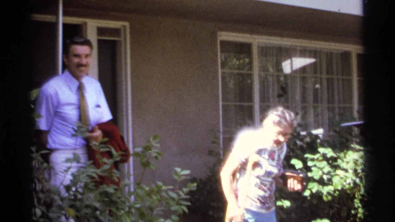 Man in light shirt and tie stands near a doorway as a woman with crutches passes shrubs.