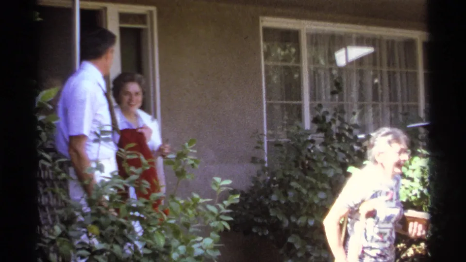 1974 La Paz home-movie frame showing adults near a doorway, including a woman using crutches in bright garden light.