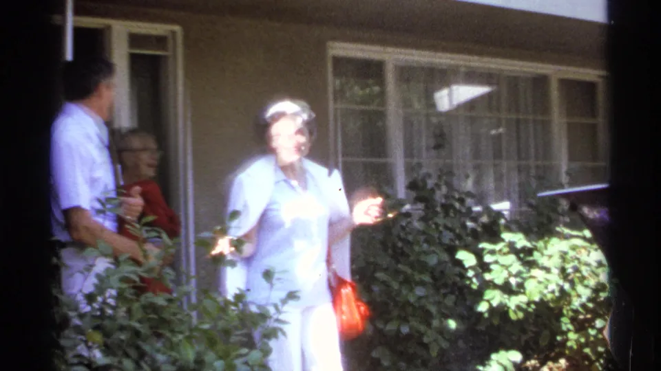 1974 La Paz frame showing a woman in a light blue outfit carrying a red bag near garden shrubs and a house entrance.