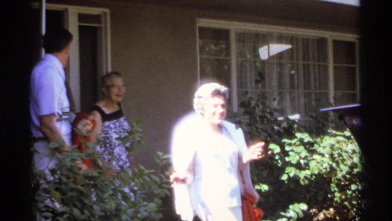 Three adults stand outside a house while an older woman in a pale dress smiles near garden bushes.