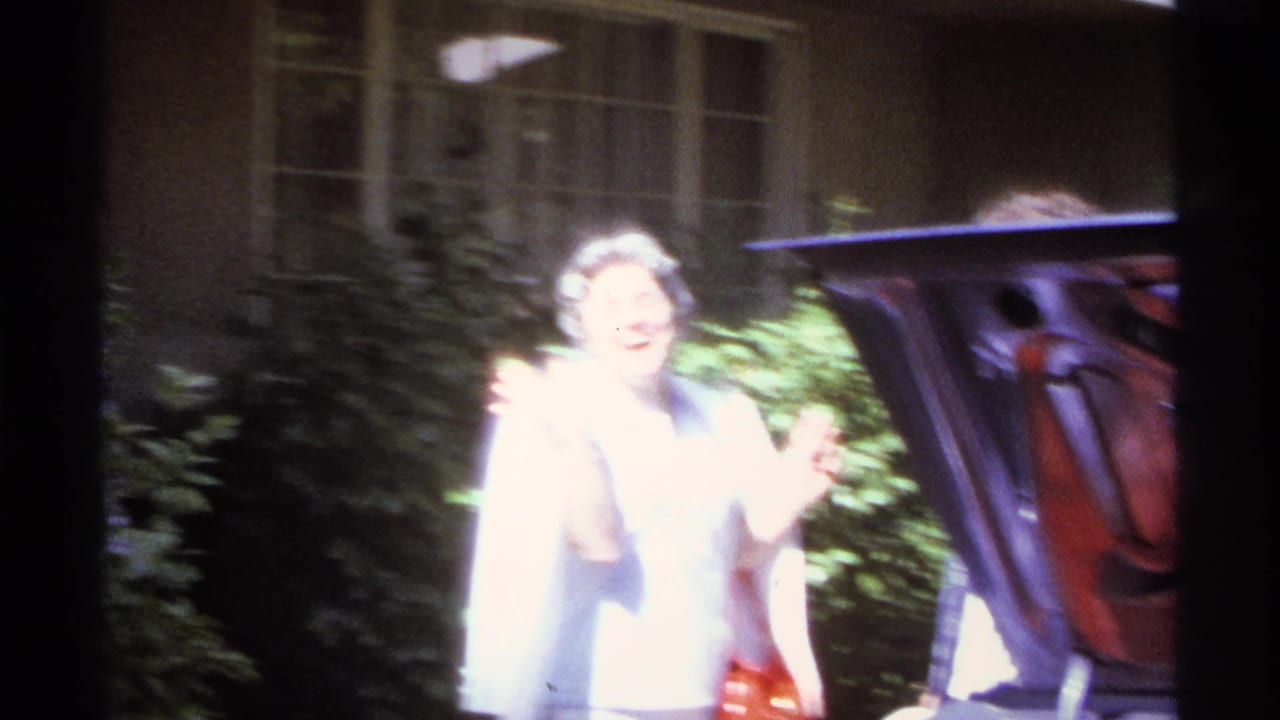 Older woman in bright sunlight gestures near an open car trunk outside a house.
