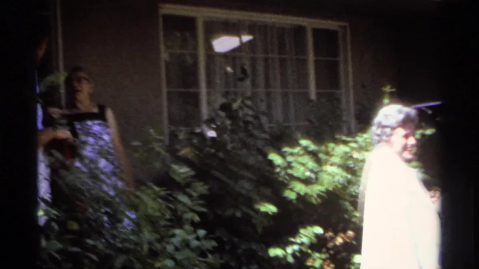The older woman turns back toward the camera in a 1974 La Paz garden scene, with another adult partly visible at left.