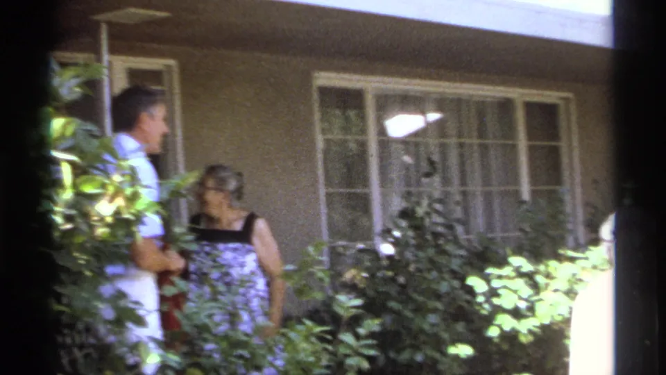 A man in a light shirt and a woman in a patterned dress talk outside a La Paz house while the older woman exits frame.