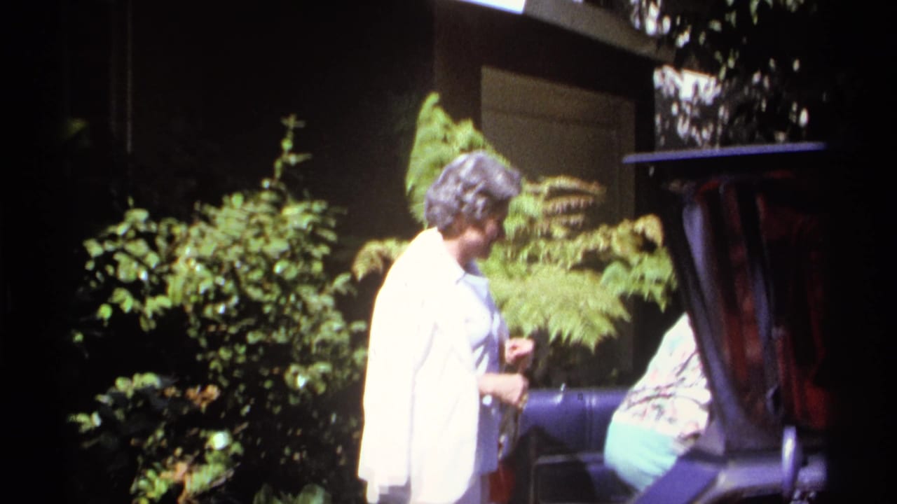 Adults near bushes and a porch, with a woman in a light top facing away.