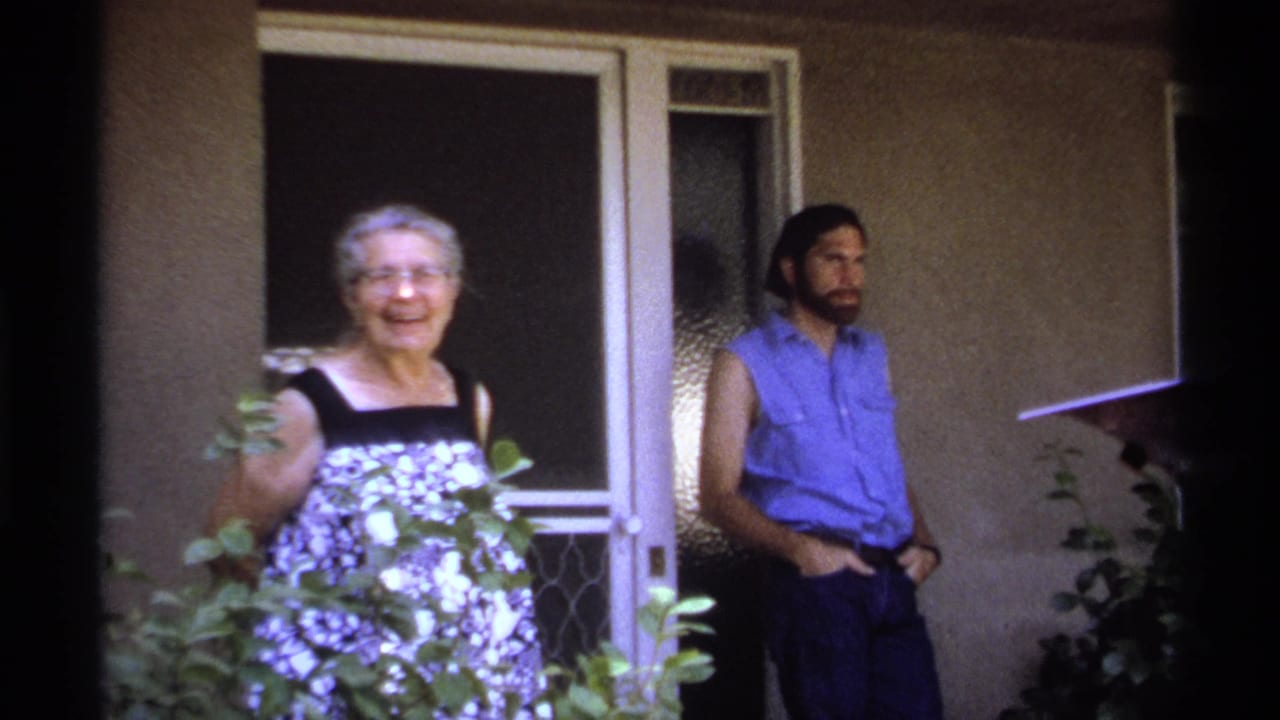 Elderly woman smiling near a doorway while a man in a blue sleeveless shirt stands beside her.