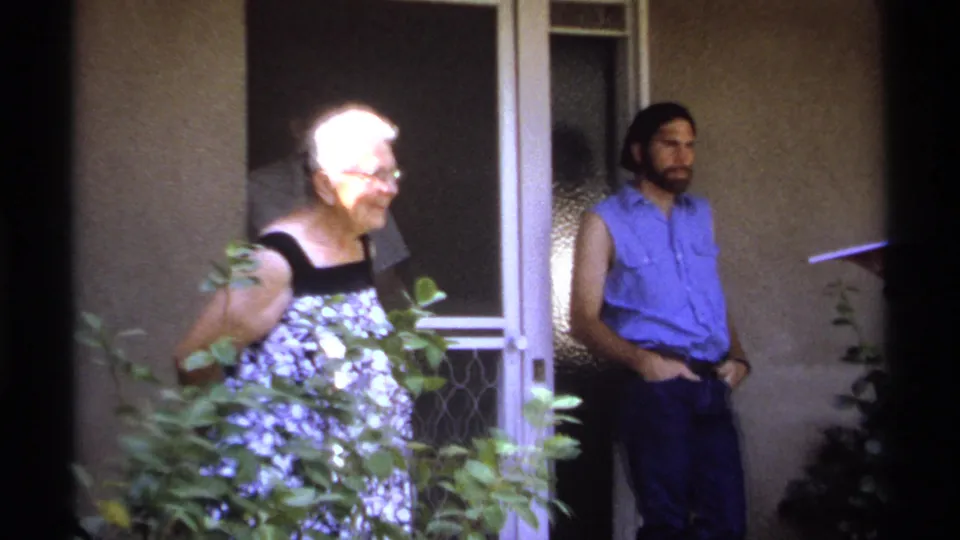 1974 La Paz porch scene with an elderly woman in glasses near bushes and a man standing by the door.