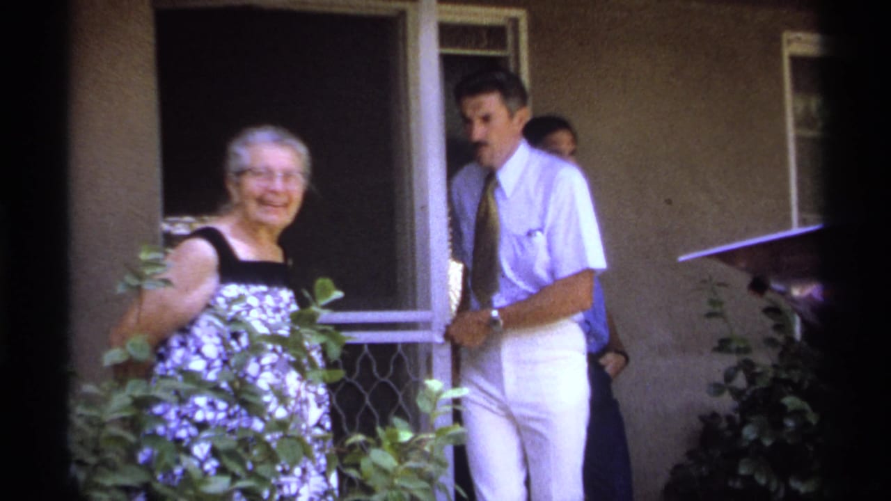 Elderly woman smiles near shrubs while a man in a tie stands by a doorway.