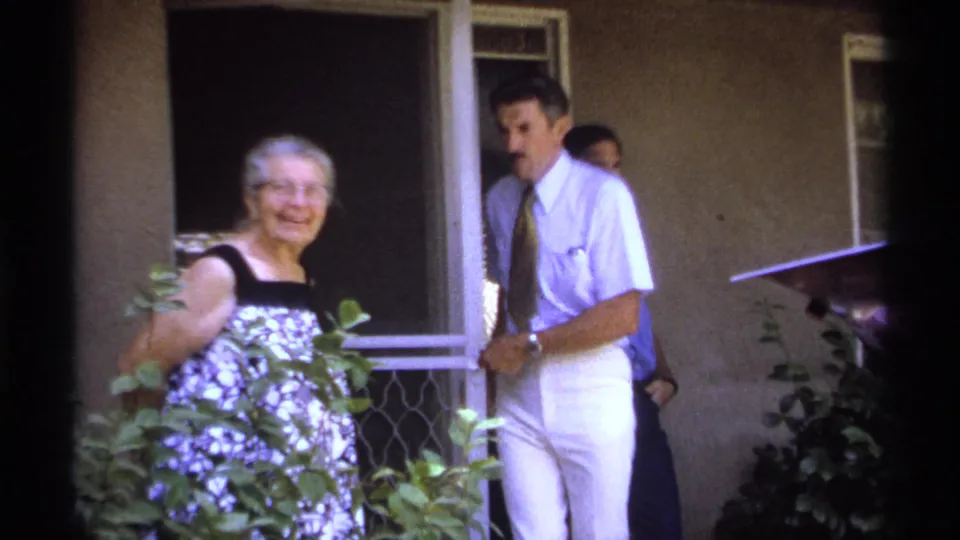 In 1974 La Paz home-movie footage, an elderly woman smiles beside porch foliage as a man in white trousers stands near a screen door.