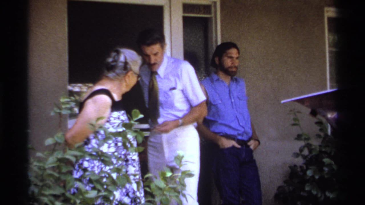 Elderly woman turns toward a man in a tie while another man stands near the wall.