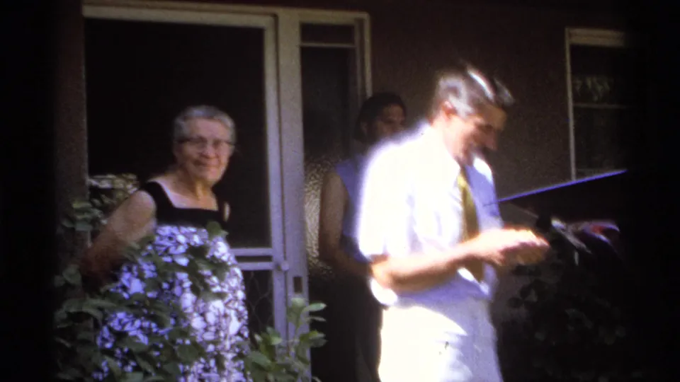 A bright 8mm frame shows the elderly woman near a porch door while the man in a tie looks down, partly overexposed by sunlight.