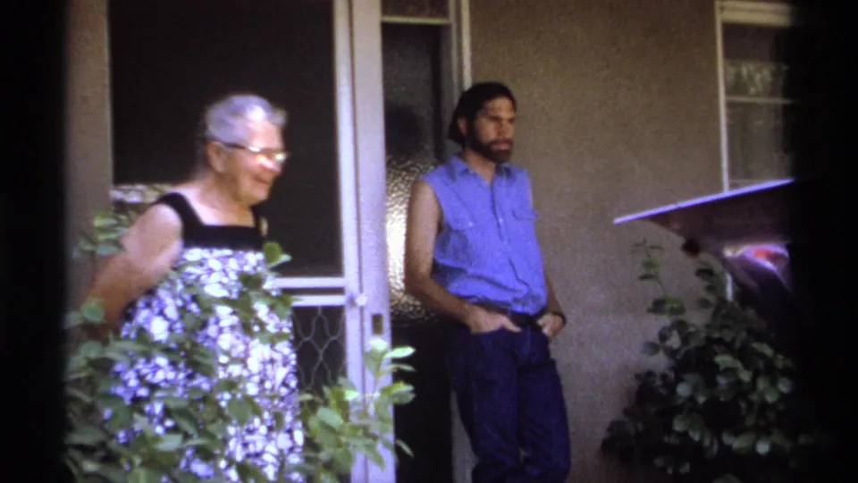 In the closing frame, the elderly woman smiles near the La Paz porch entrance as a man in a blue sleeveless shirt stands relaxed against the wall.