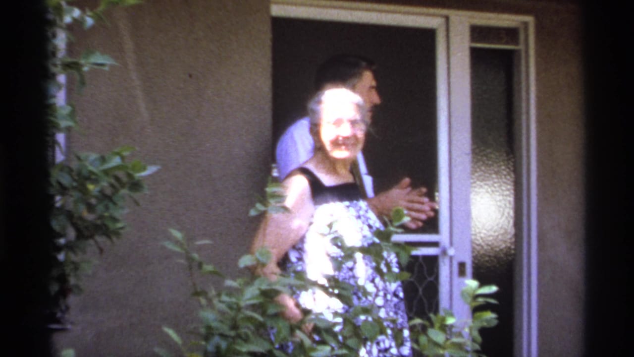 Older woman smiling near a doorway with a man standing behind her.