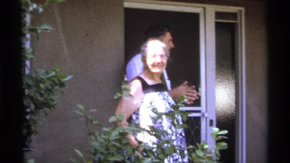 In 1974 La Paz home-movie footage, an older woman in a patterned dress smiles near a doorway as a man stands behind her.
