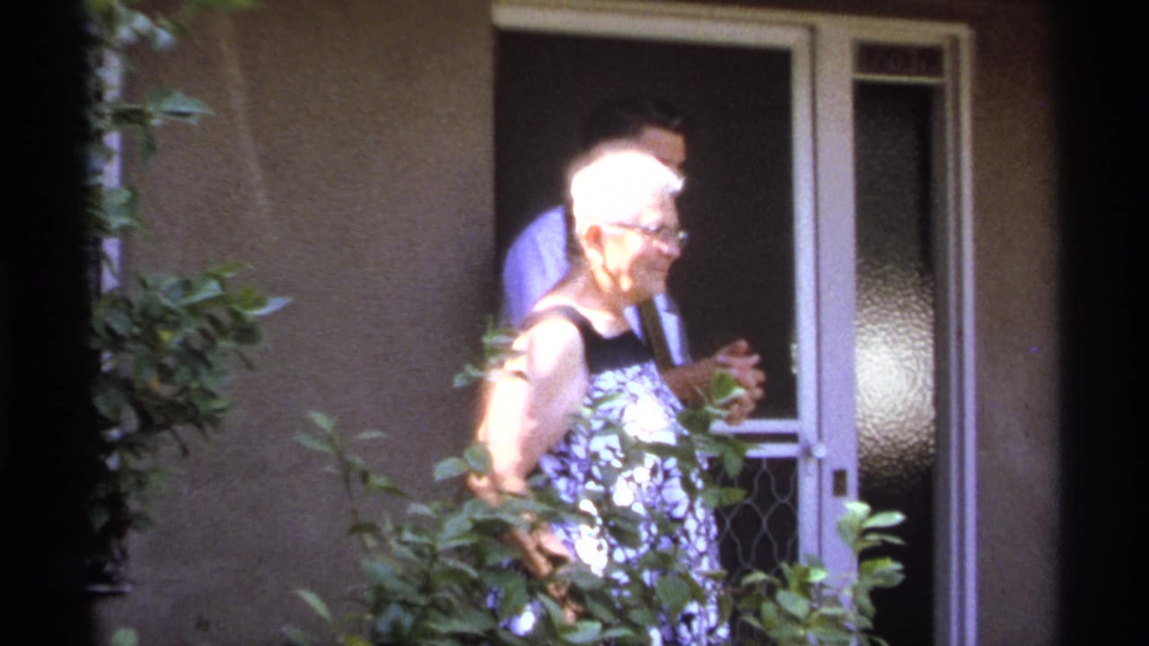 Older woman in profile beside a doorway with a man partly visible behind her.