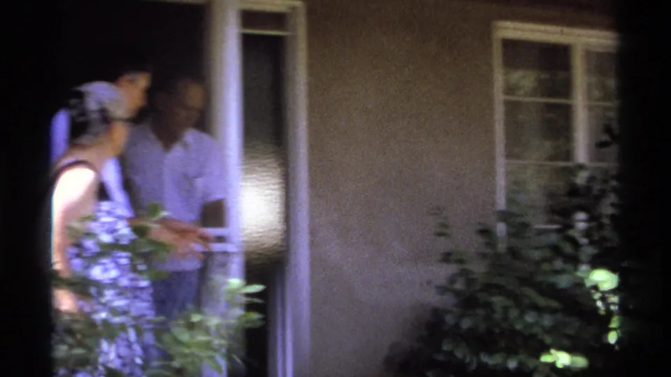 The older woman and a man stand near the doorway, partly hidden by garden leaves and deep film shadow in the La Paz home movie.