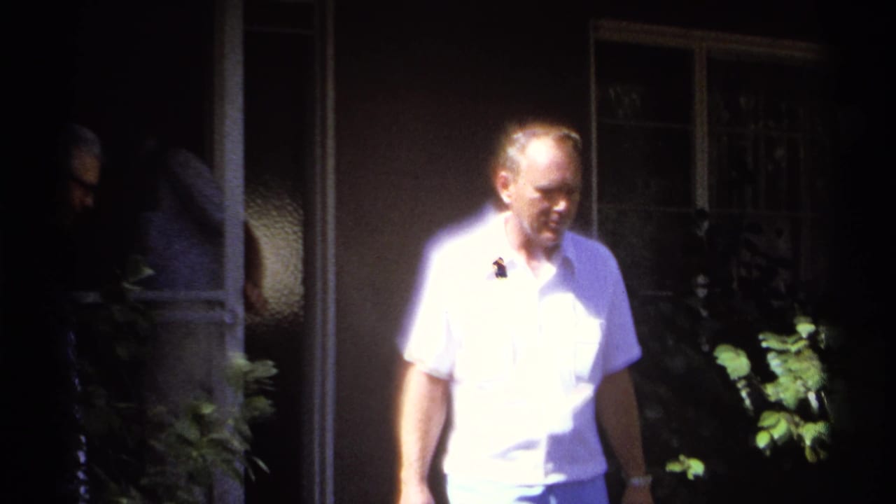 A man in a white short-sleeve shirt walks in bright sunlight outside a house.