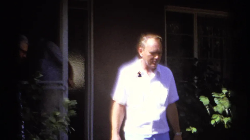 A man in a white shirt walks past shaded windows and foliage in 1974 La Paz home-movie footage.