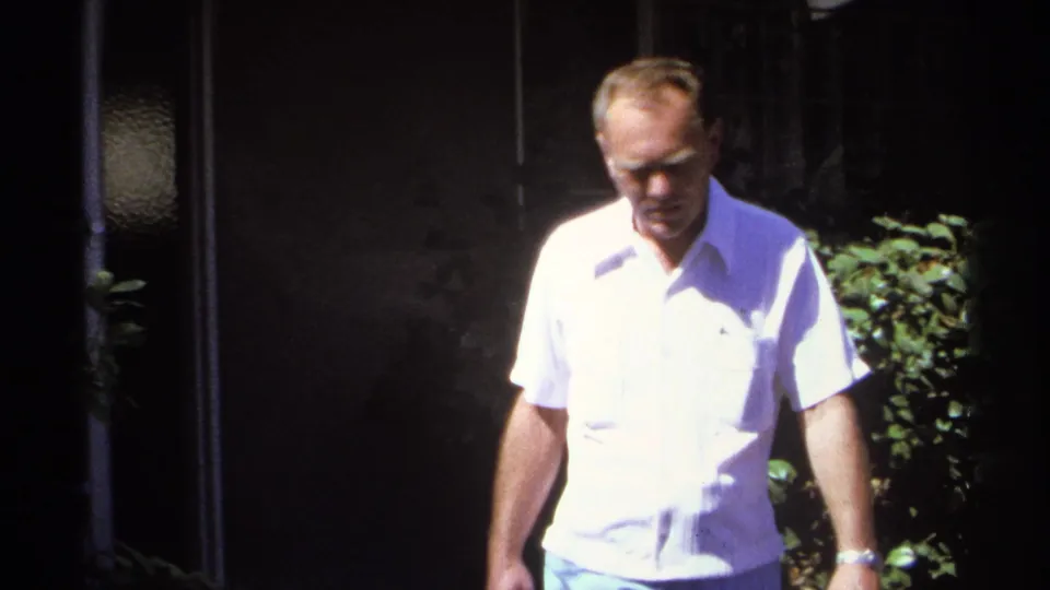 The man walks closer to the 8mm camera, with shrubs and a shaded house exterior behind him in La Paz, Mexico.
