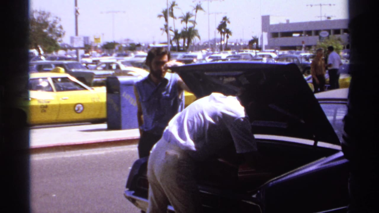 In La Paz, Mexico, 1974, men stand near an open car trunk in a bright parking lot with parked cars, palm trees, and a yellow vehicle behind them.