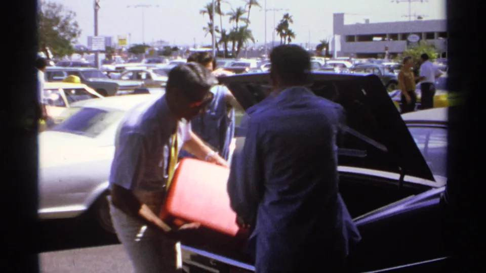 A man in a light shirt removes a red suitcase from an open car trunk in a La Paz parking lot, with another man and vintage cars nearby.