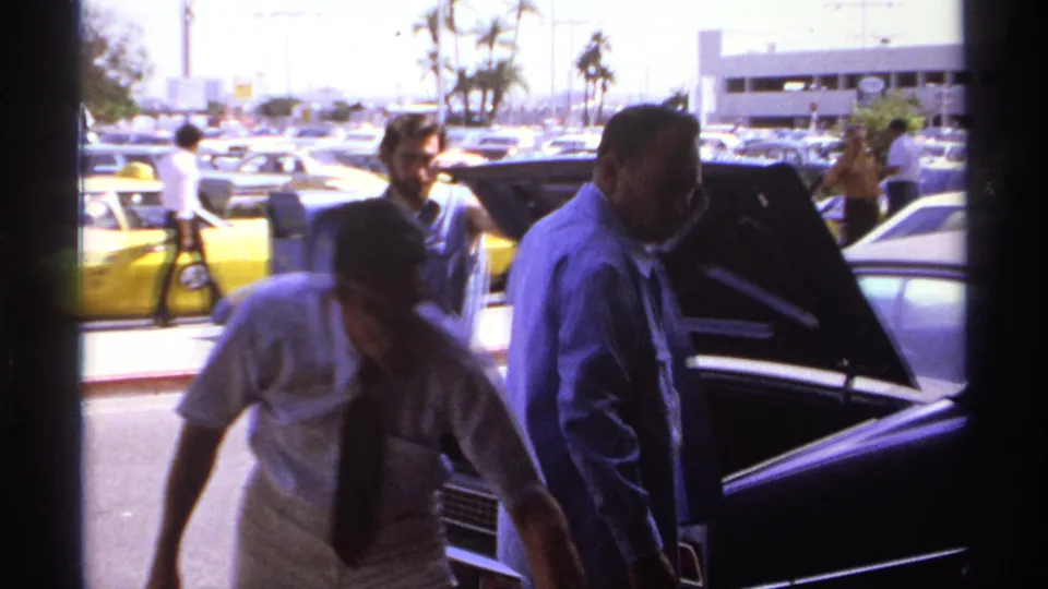 People pass around an open car trunk in a bright La Paz parking lot, with 1970s cars, palm trees, and a low building visible behind them.