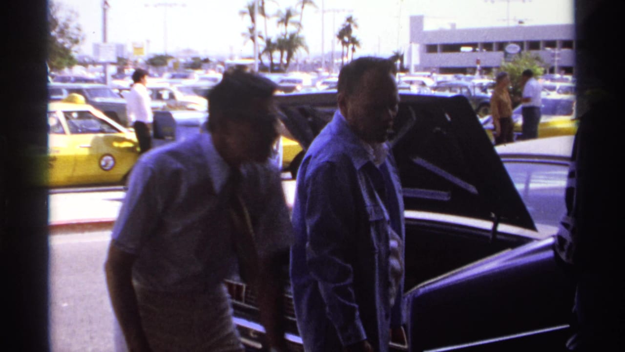Men stand near an open dark sedan trunk with yellow taxis and parked cars behind them.