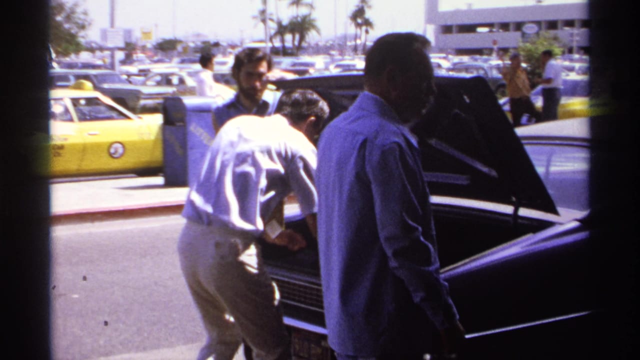 A man in a white shirt bends into an open car trunk while other men stand nearby.