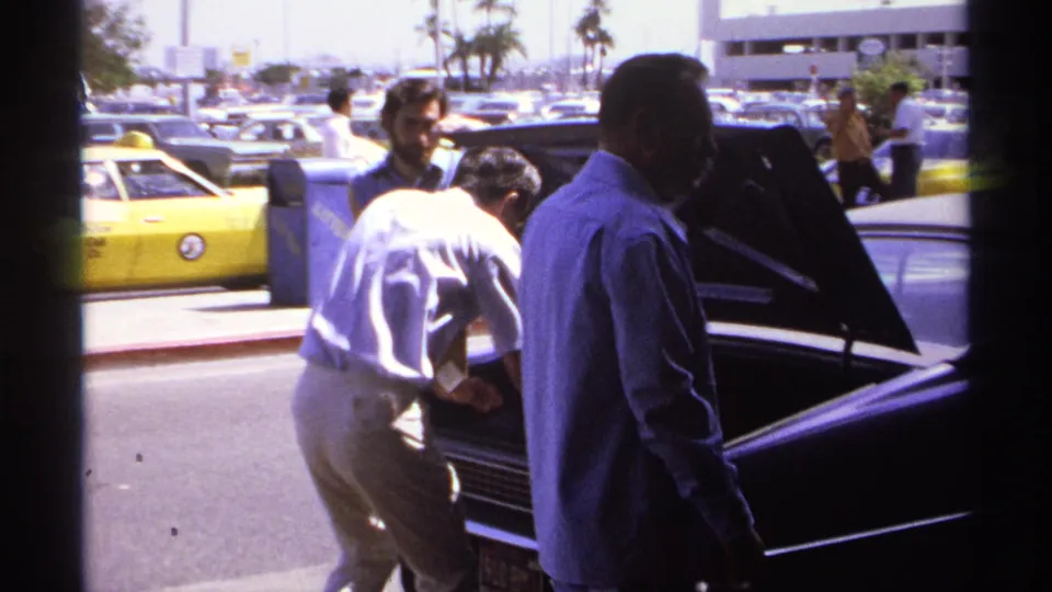 In this 1974 La Paz frame, a man in a white shirt reaches into the open trunk of a dark sedan as others watch nearby.