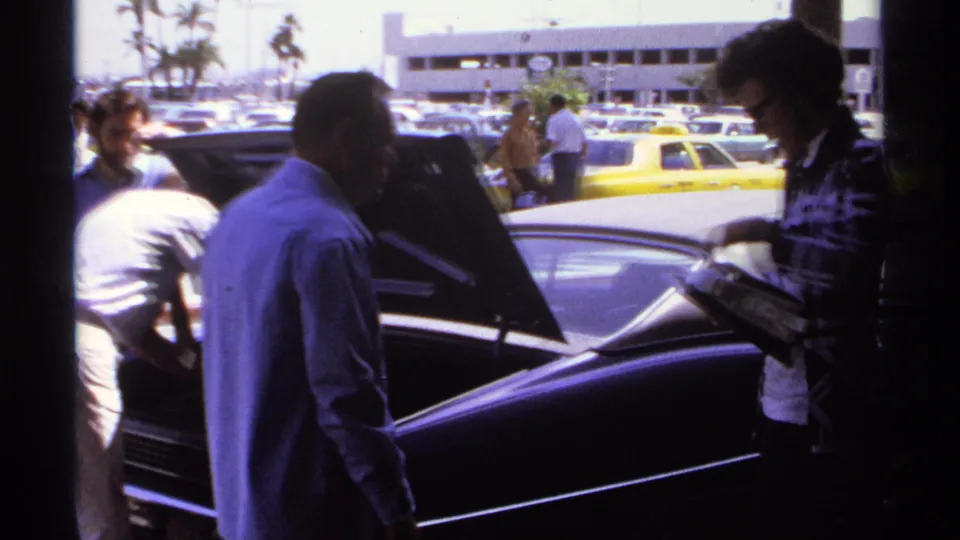 1974 La Paz footage frame with men around a dark sedan's open trunk, yellow taxis, cars, and palm trees in the background.