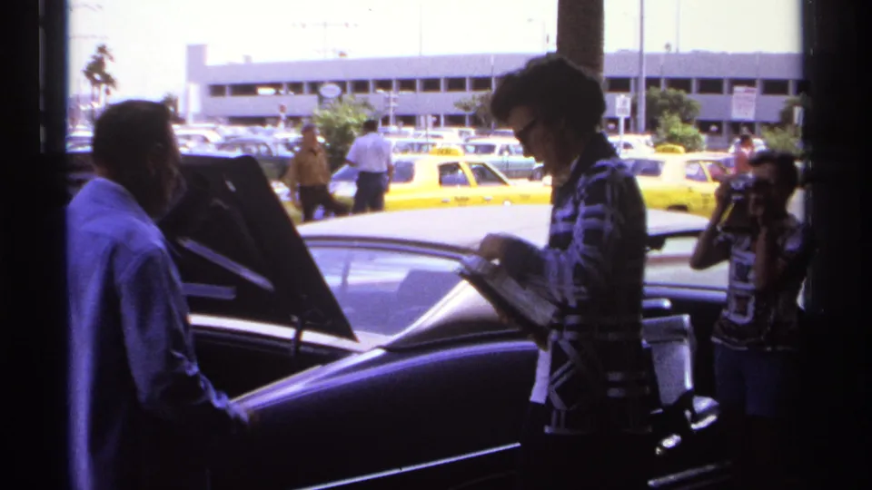 Final 1974 La Paz frame showing a man handling an item near the open trunk as other people and taxis appear in the parking lot.