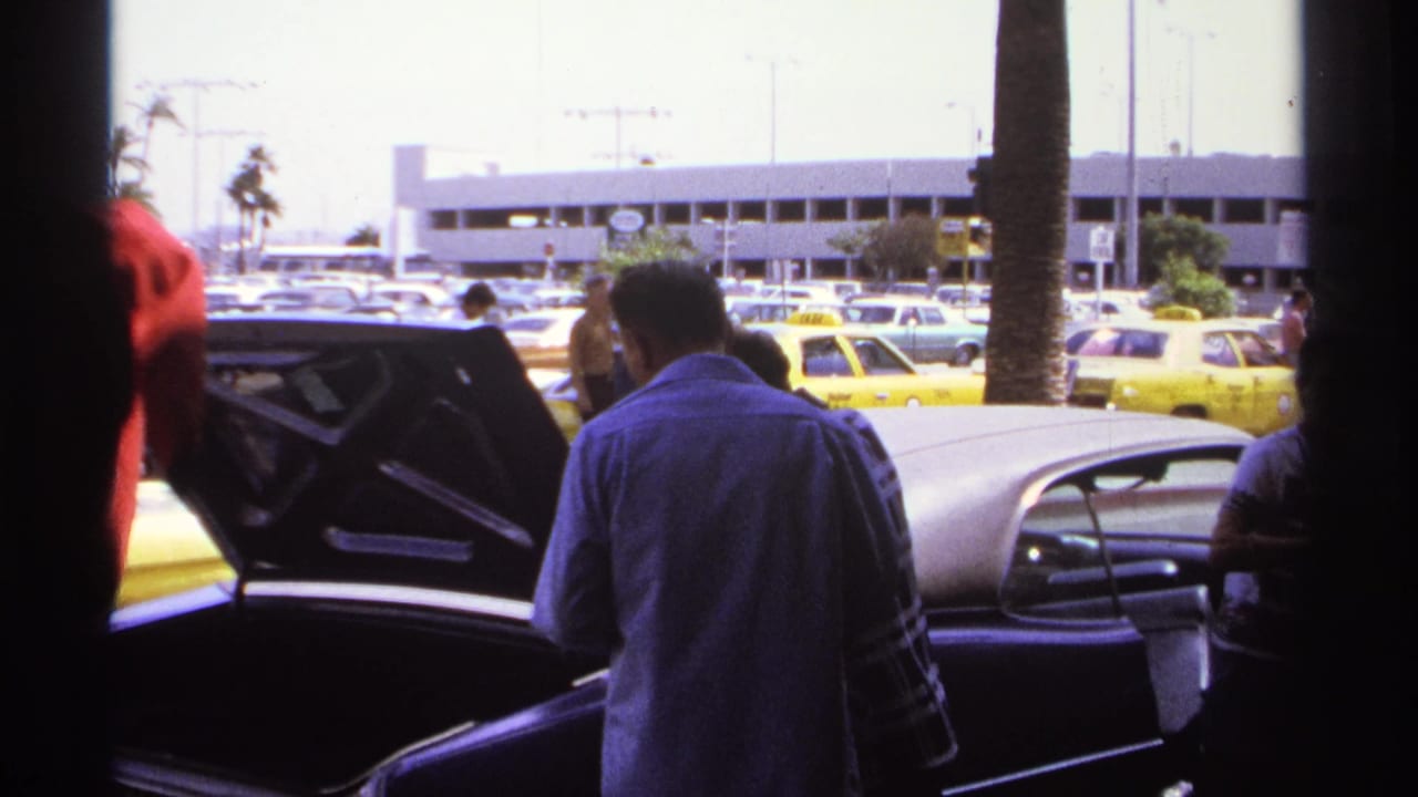 Adults stand beside a dark car with its trunk open in a sunny parking area.