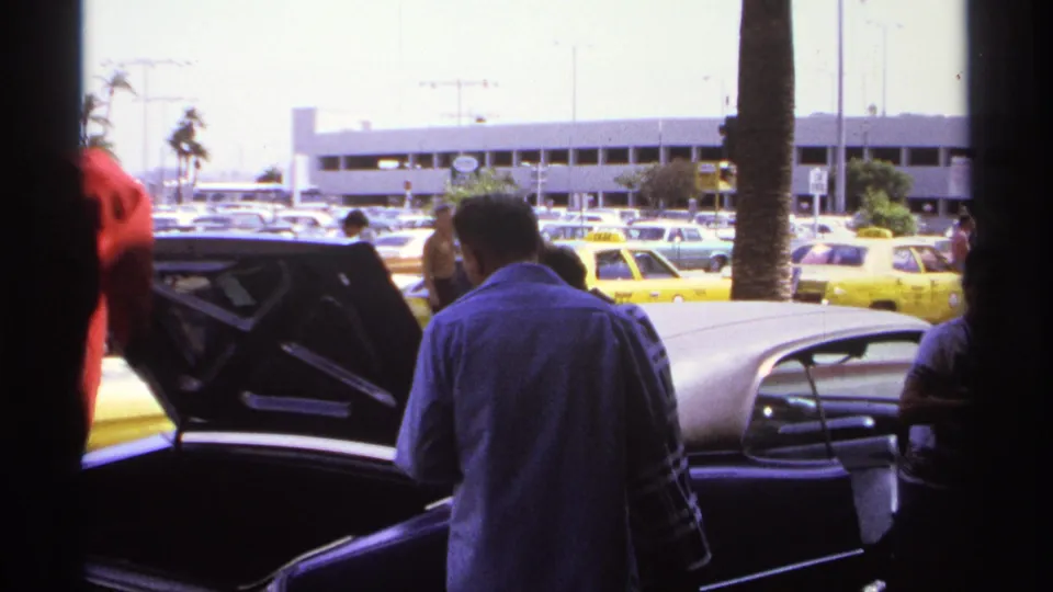 1974 La Paz home-movie frame showing adults near an open car trunk with yellow taxis and parked cars behind them.