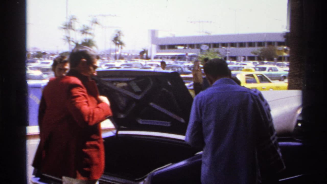 A man in a red jacket stands near an open car trunk while another man faces the car.