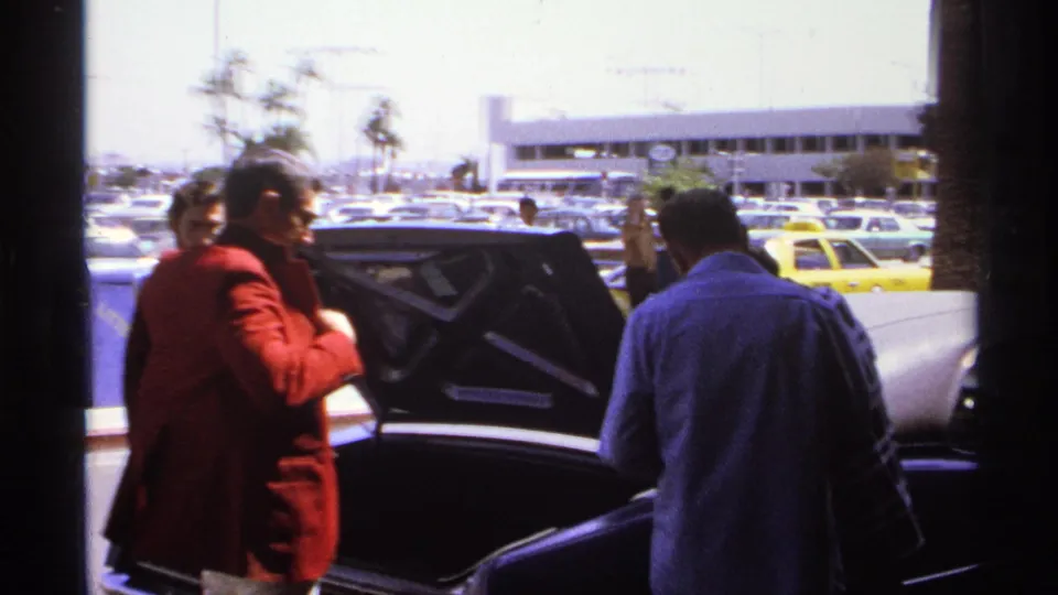 1974 La Paz frame with a man in a red jacket beside an open trunk, another man in blue, taxis, palms, and parked cars.