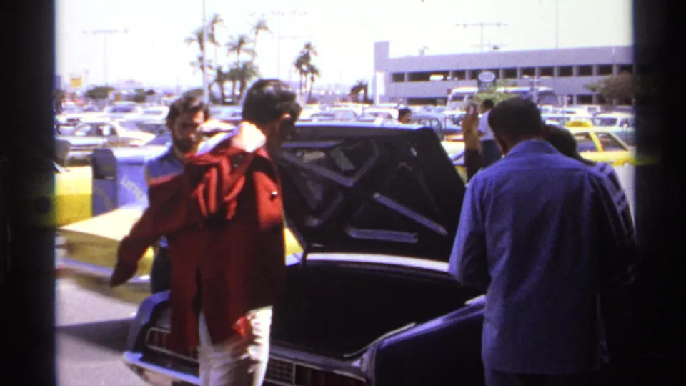 1974 La Paz home-movie frame showing a man adjusting a red jacket near an open car trunk in a busy parking area.