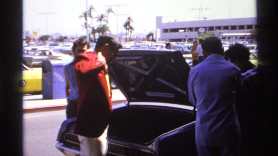 1974 La Paz frame of a man adjusting his red jacket next to an open car trunk, with adults and taxis in the background.