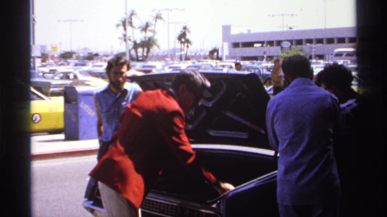 Men stand by an open car trunk at a sunny airport curbside.