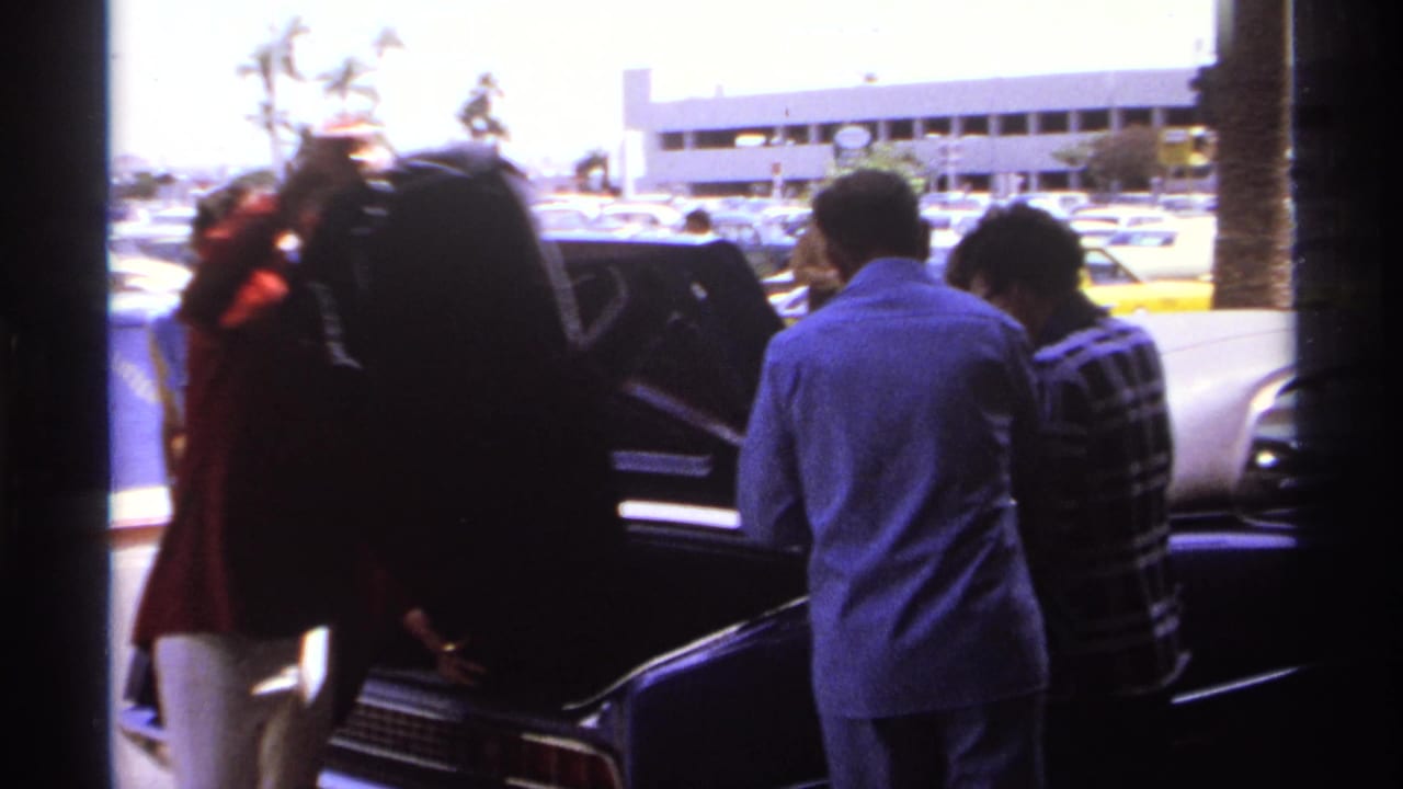 Several men gather around an open trunk beside parked airport cars.