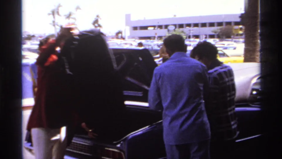 Men at a La Paz airport curb gather around an open car trunk, with parked cars and palm trees behind them.